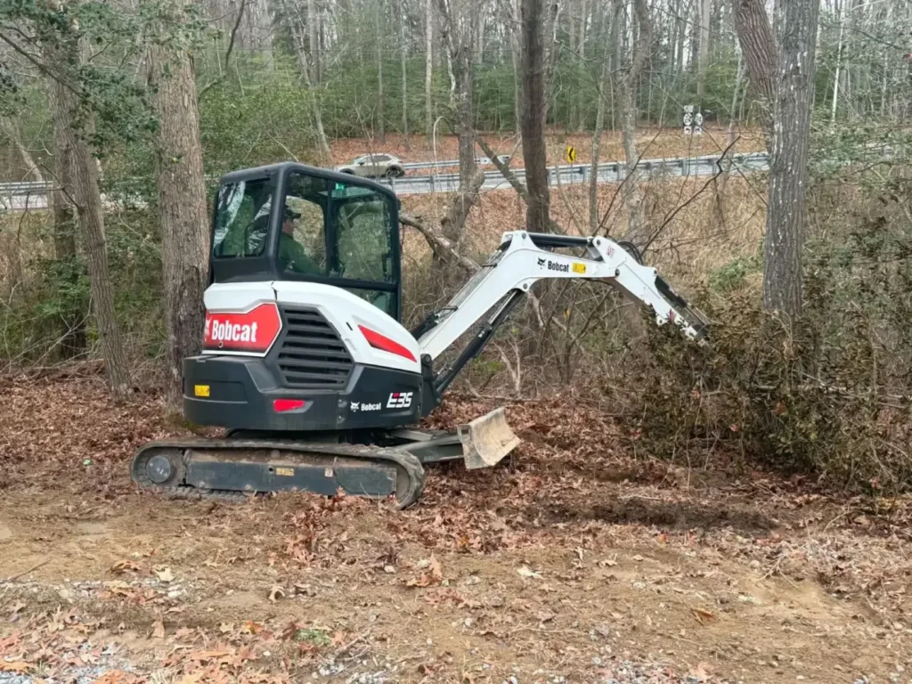 Mini excavator pulling brush at the edge of a wooded lot